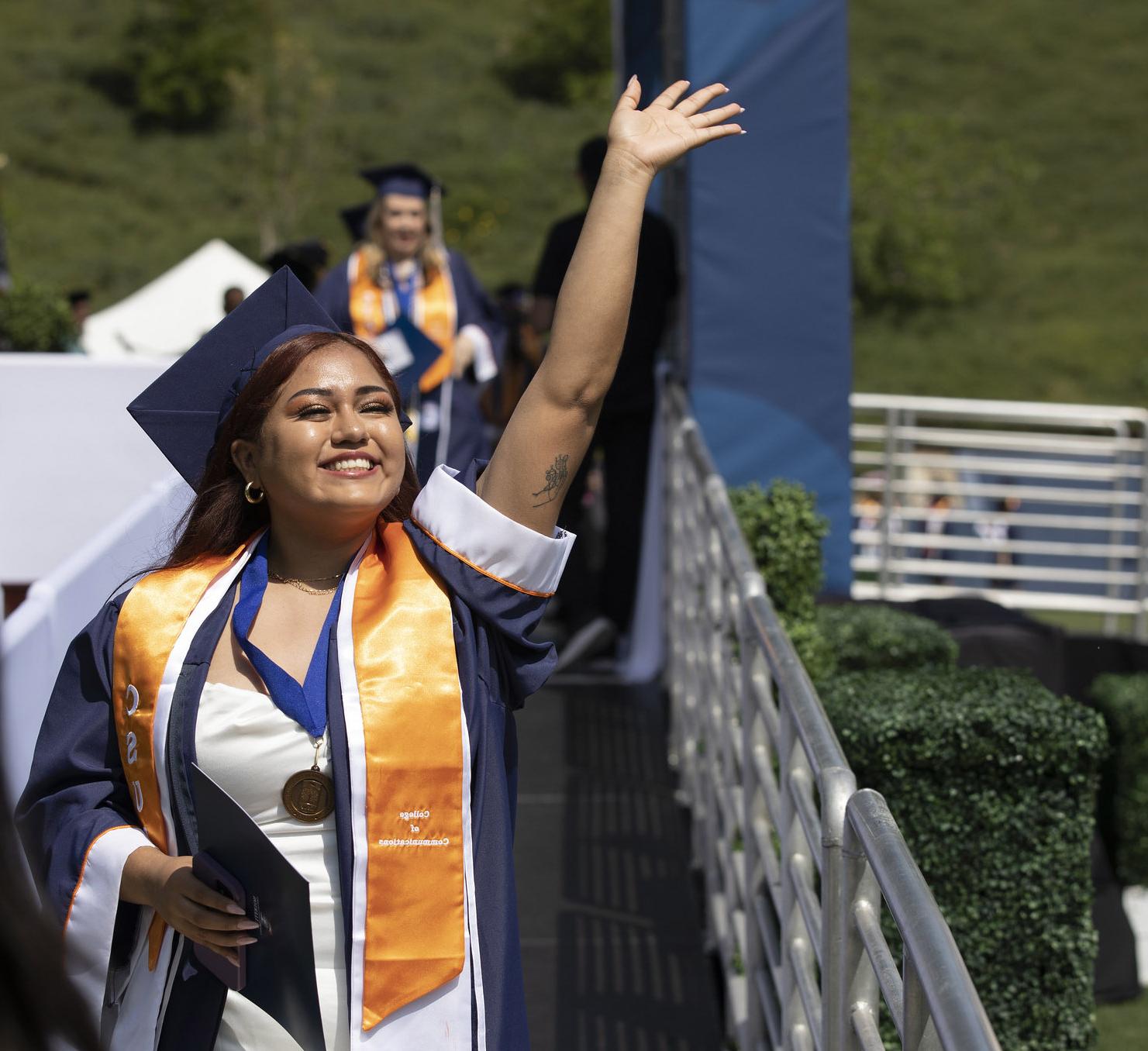Latina student waves at crowd as she walks offstage after receiving her diploma during commencement.